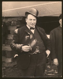Lodret, sort / hvidt fotografi af en mand i en derbyhue og sløjfe, der ryger en cigar
